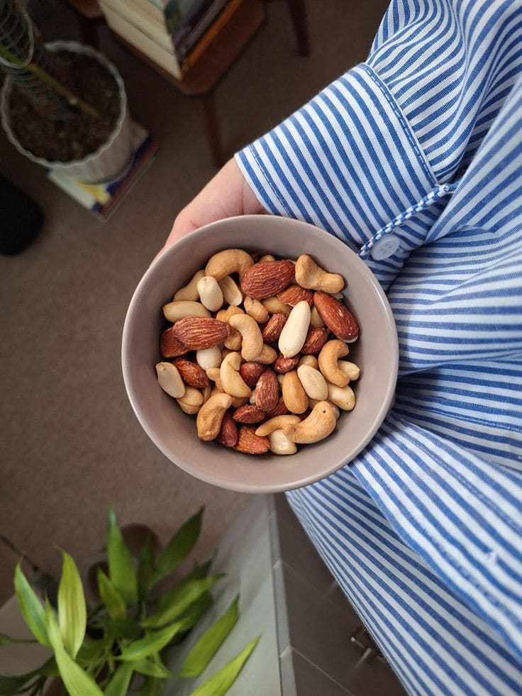 A wooden bowl of mixed nuts including almonds and cashews surrounded by pumpkin seeds on a rustic surface